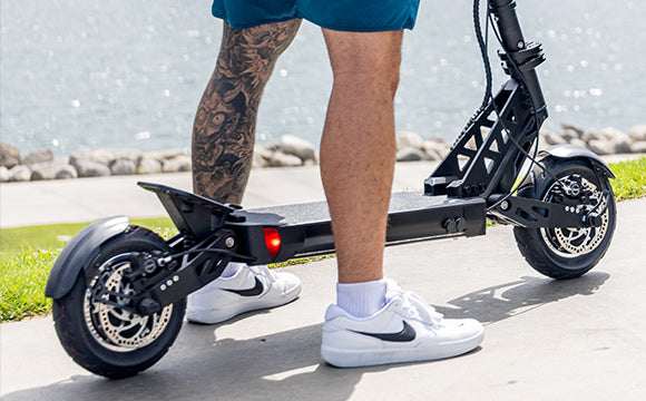 Tattooed legs straddling an electric scooter next to the beach on sidewalk